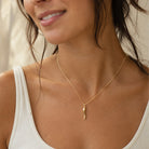 A close-up lifestyle shot of a woman wearing a delicate gold chain necklace with a small, stylized gold pendant. She is wearing a white ribbed tank top, and the background is a soft, warm beige.