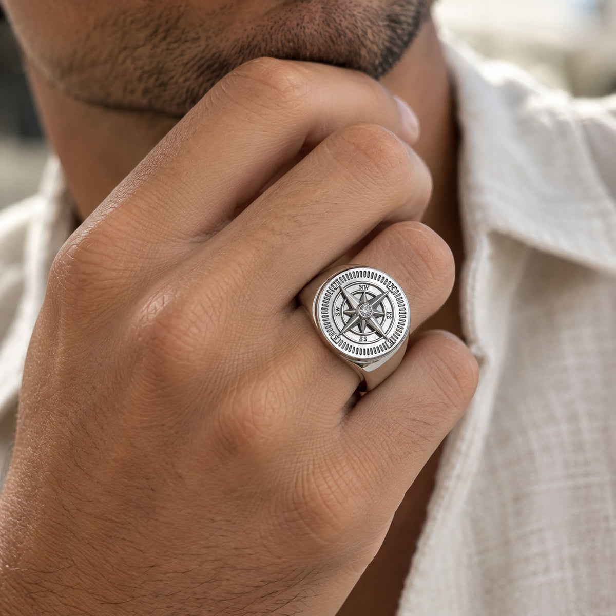 A close-up of a man's hand wearing a silver-toned signet ring with an engraved compass design, paired with a white textured shirt.