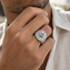 A close-up of a man's hand wearing a silver-toned signet ring with an engraved compass design, paired with a white textured shirt.