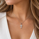 A close-up of a woman wearing a delicate silver cross pendant necklace with clear stones, styled with a white ribbed knit top.