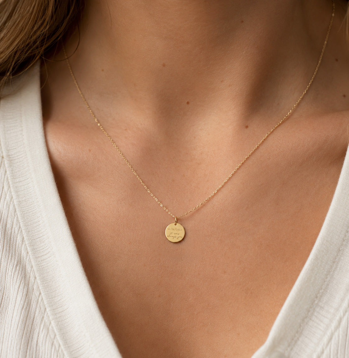[group: 10k-gold] A delicate gold chain necklace with a small round engraved disc pendant, worn on a model's neck over a white ribbed top.