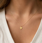 [group: 10k-gold] A delicate gold chain necklace with a small round engraved disc pendant, worn on a model's neck over a white ribbed top.