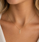 A close-up of a delicate gold chain necklace worn by a model, featuring a small vertical bar pendant with engraved text, set against warm skin tones and a white ribbed top.
