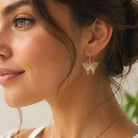 A close-up of a woman wearing a delicate gold butterfly drop earring with a white center, paired with a thin gold chain necklace.