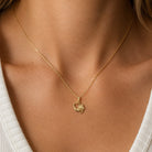 A close-up of a delicate gold chain necklace worn by a model, featuring a small heart-shaped pendant with a floral design, paired with a white ribbed top.