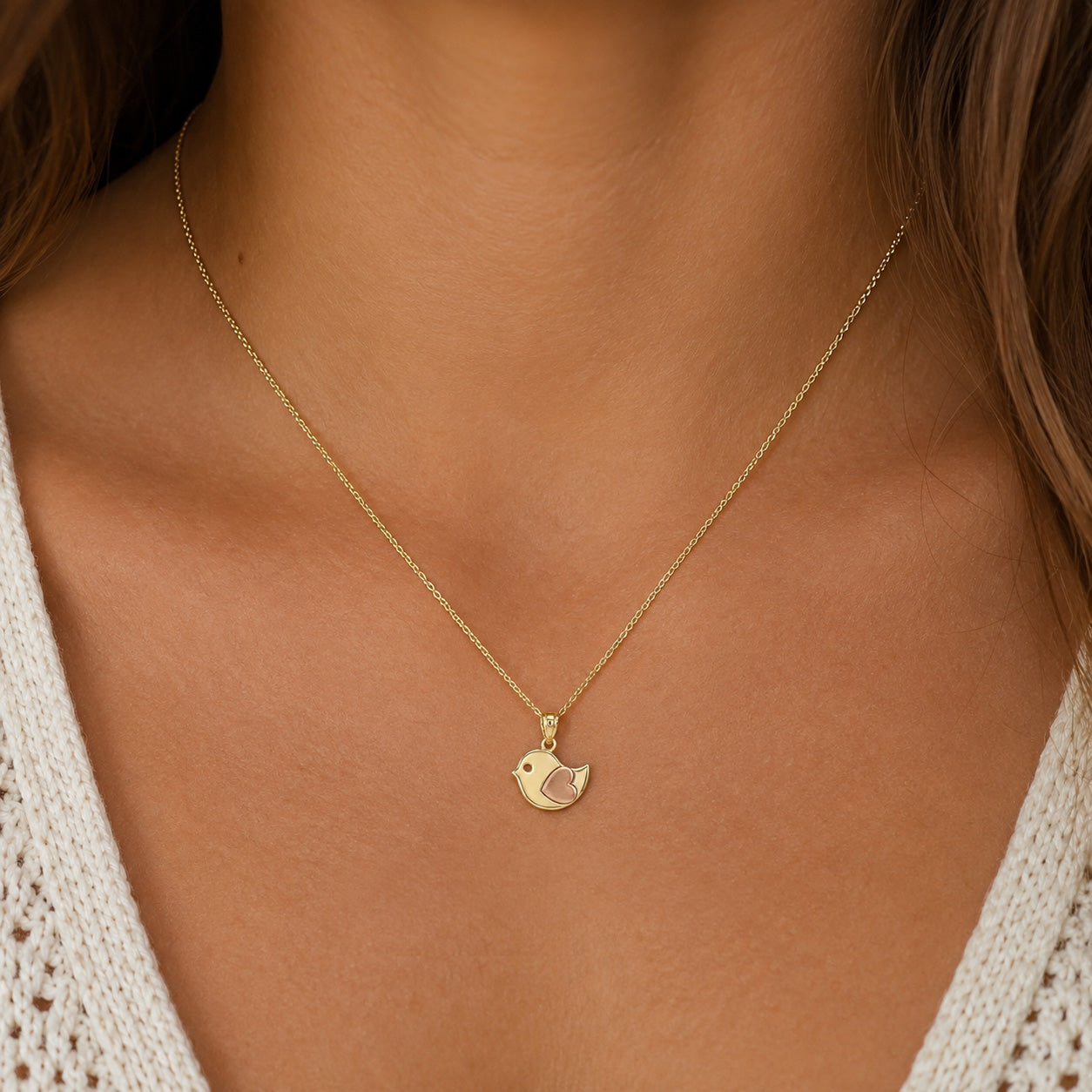 A delicate gold chain necklace with a small bird-shaped pendant, worn on the neck of a model wearing a white knit top.