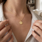 Gold necklace with a pendant worn by a person, with a blurred indoor background.