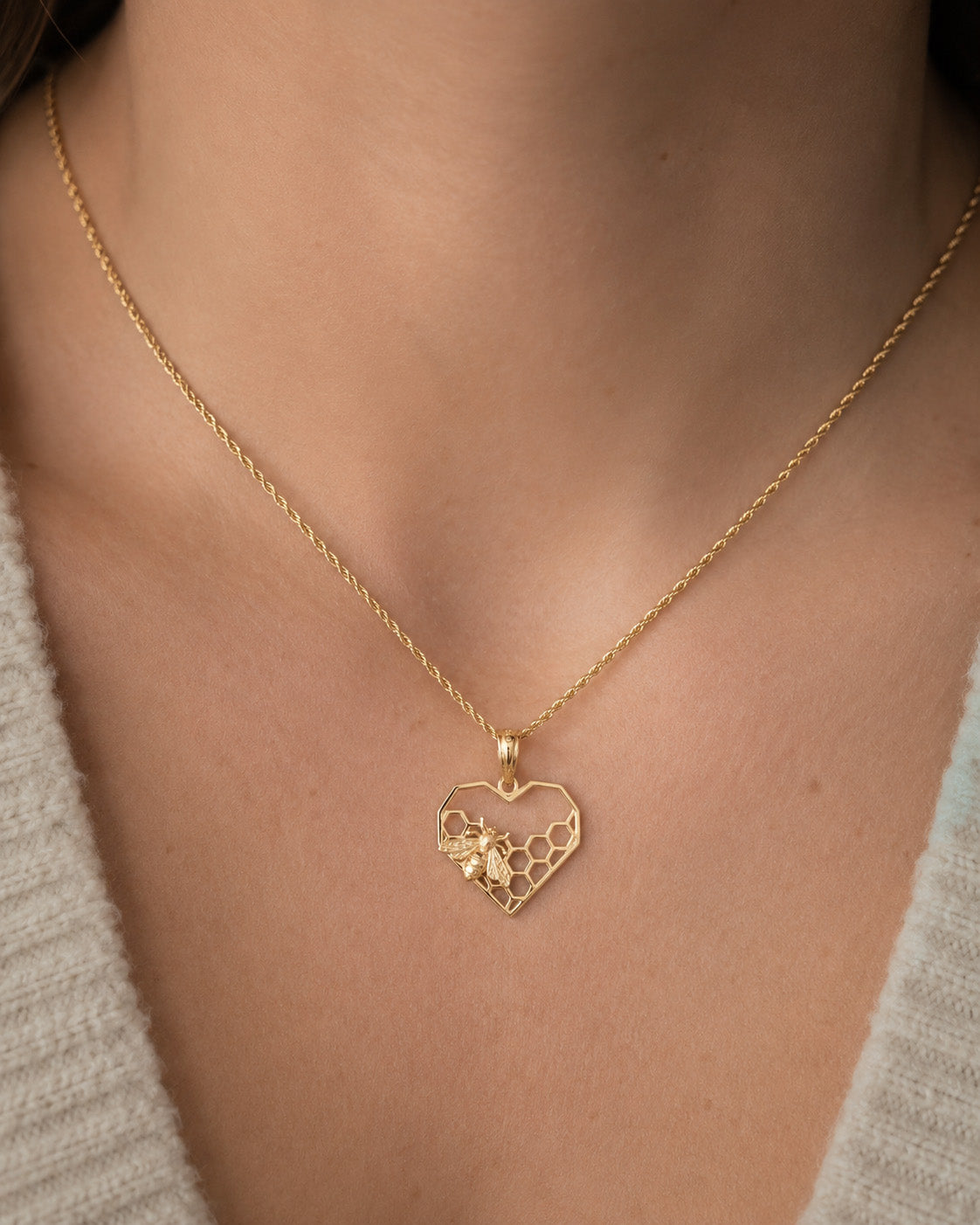 A close-up of a person wearing a delicate gold chain necklace with a heart-shaped pendant featuring a honeycomb pattern and a small flower detail.