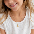 A close-up lifestyle shot of a young girl wearing a delicate gold chain necklace with a small gold bunny pendant, paired with a white eyelet top.