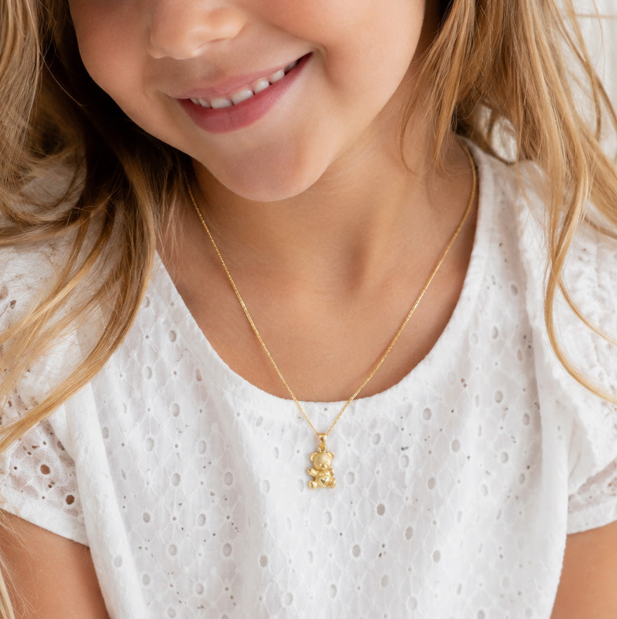 A close-up lifestyle shot of a young girl wearing a delicate gold chain necklace with a small gold bunny pendant, paired with a white eyelet top.