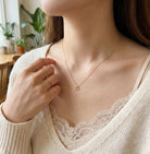 Close-up of a person wearing a delicate gold necklace and rings, with a blurred indoor background.