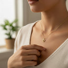 A close-up lifestyle shot of a woman wearing a delicate gold heart pendant necklace, adjusting it with her hand. She is wearing a soft cream-colored top, and a blurred indoor background with a plant is visible in the background.