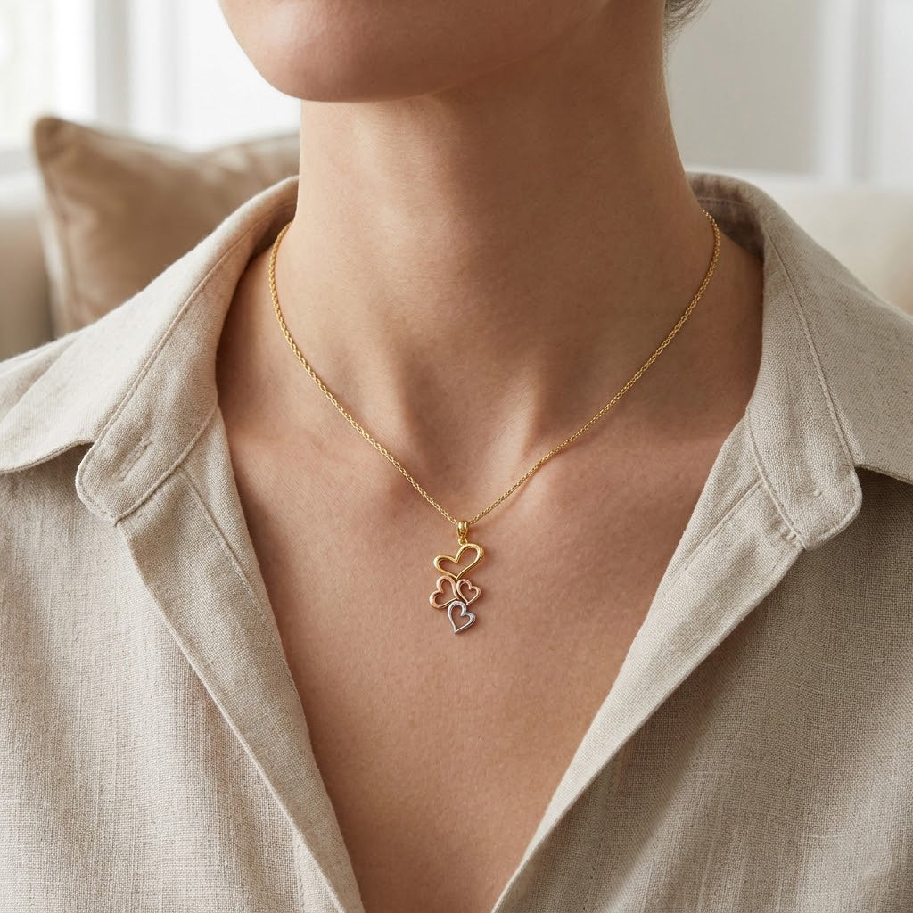 A close-up of a model wearing a delicate gold chain necklace with a multi-heart pendant in gold, rose gold, and silver, styled with a beige linen shirt.