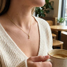 [group: d-10k-gold]A woman wearing a delicate gold necklace with a small pendant, holding a ceramic coffee cup in a cozy cafe setting.