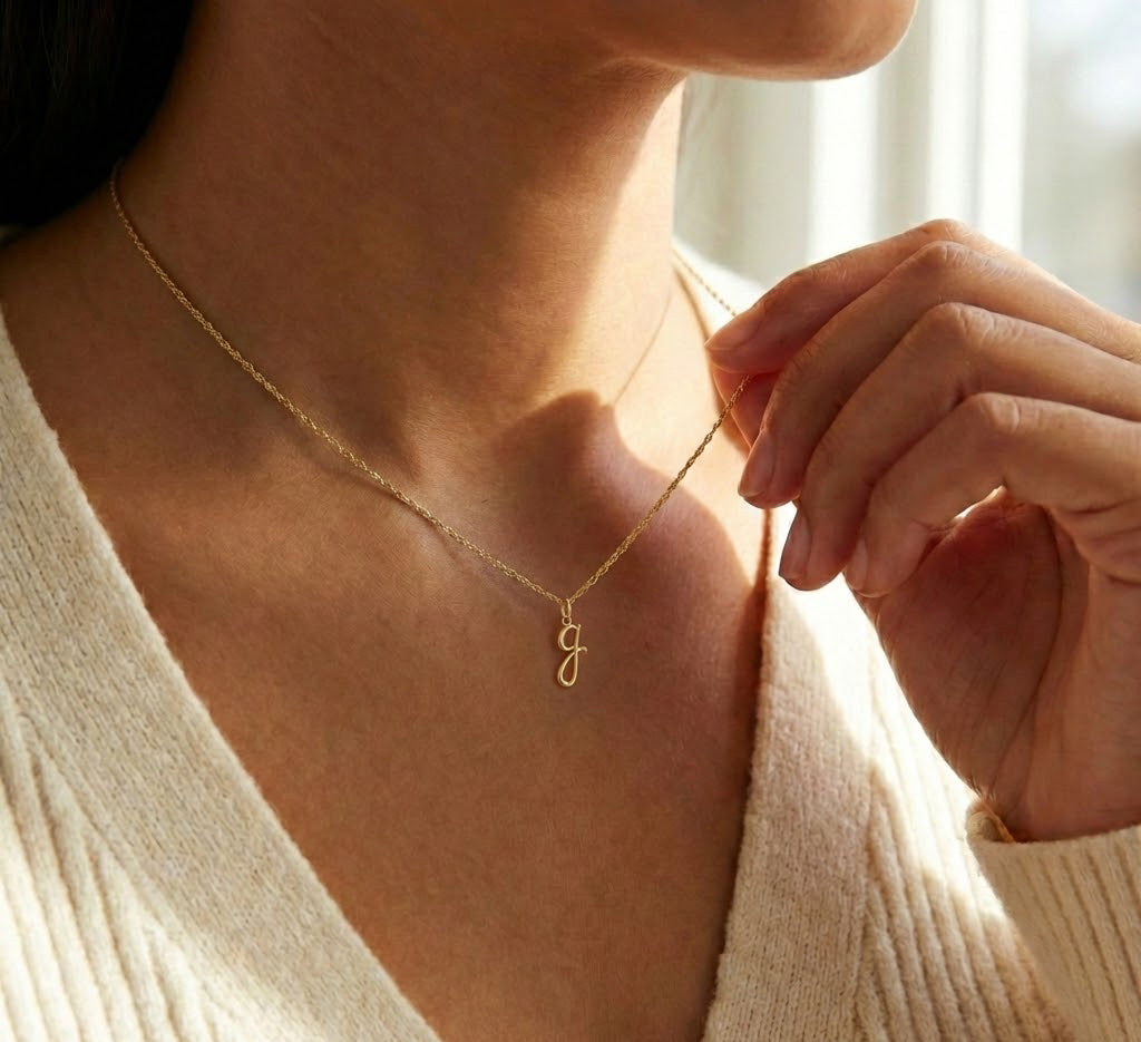 [group: g-10k-gold] A close-up lifestyle shot of a model wearing a delicate gold chain necklace with a small cursive initial 'G' pendant, styled with a cream ribbed knit top.