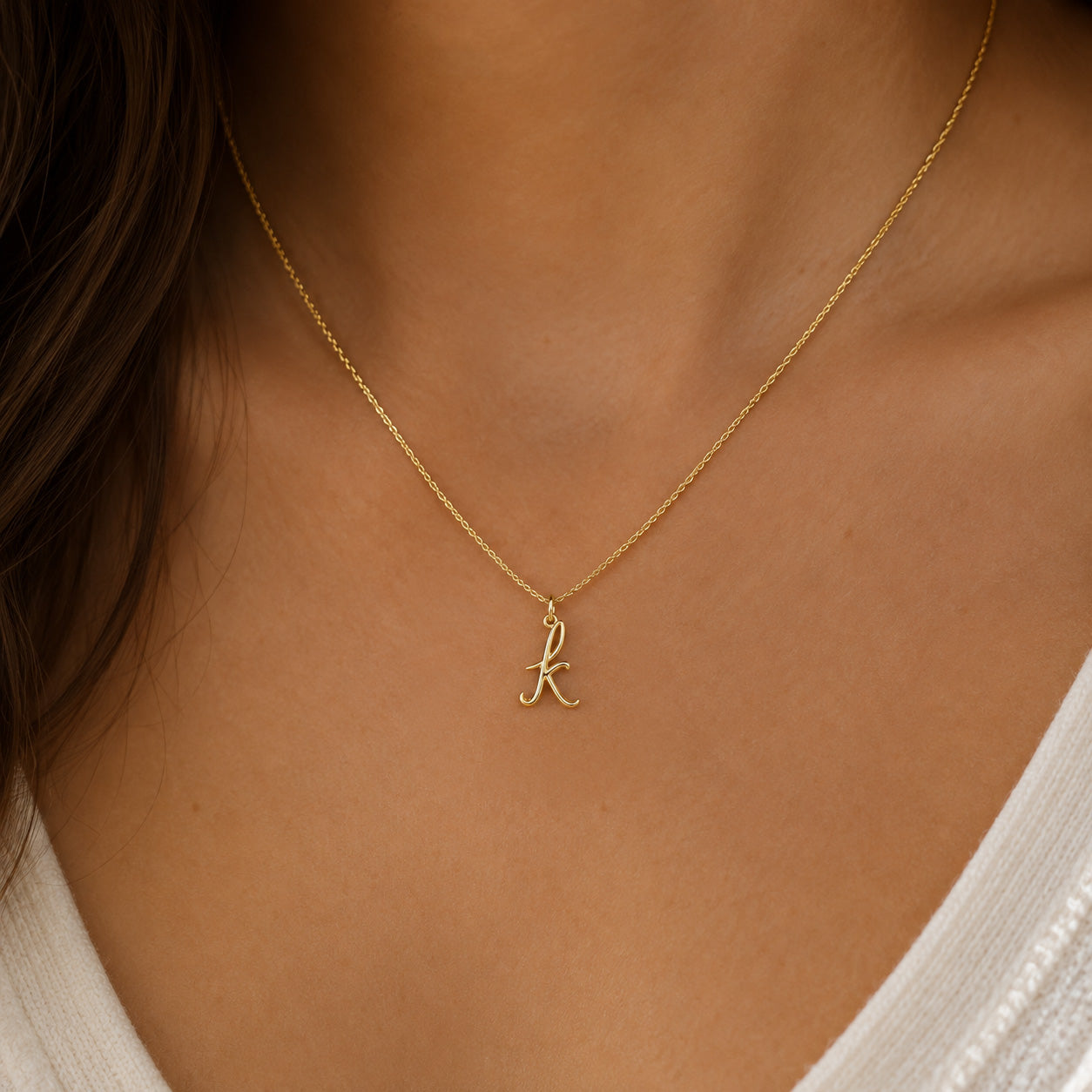 [group: k-10k-gold]A delicate gold chain necklace with a small cursive letter 'k' pendant, worn on a model's neck over a white top.