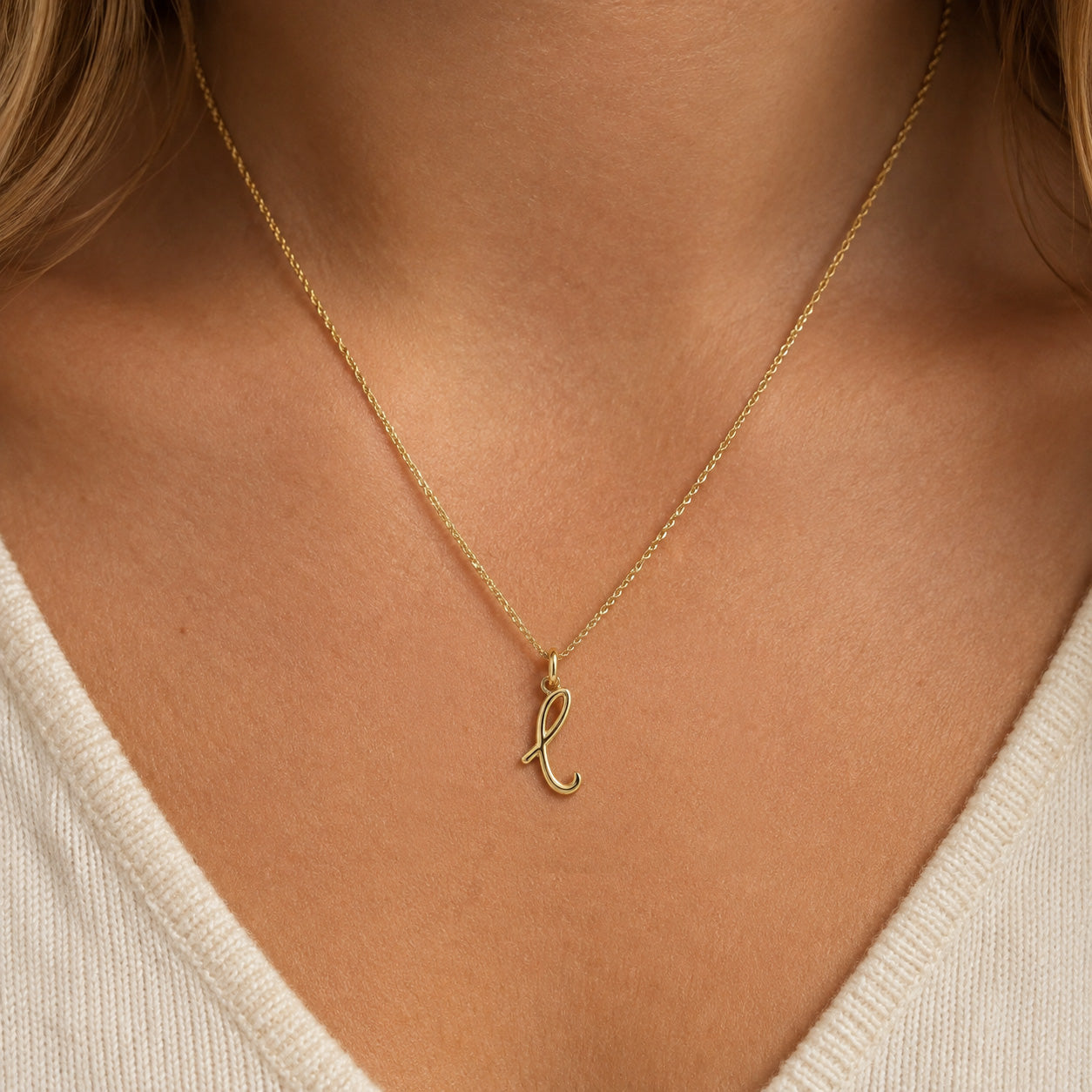 [group: l-10k-gold]A delicate gold chain necklace with a small cursive initial pendant, shown worn on a model's neck over a cream-colored V-neck top.