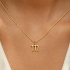 [group: m-10k-gold]A close-up of a person's neck and chest wearing a delicate gold chain necklace with a zodiac sign pendant, specifically the Libra symbol.