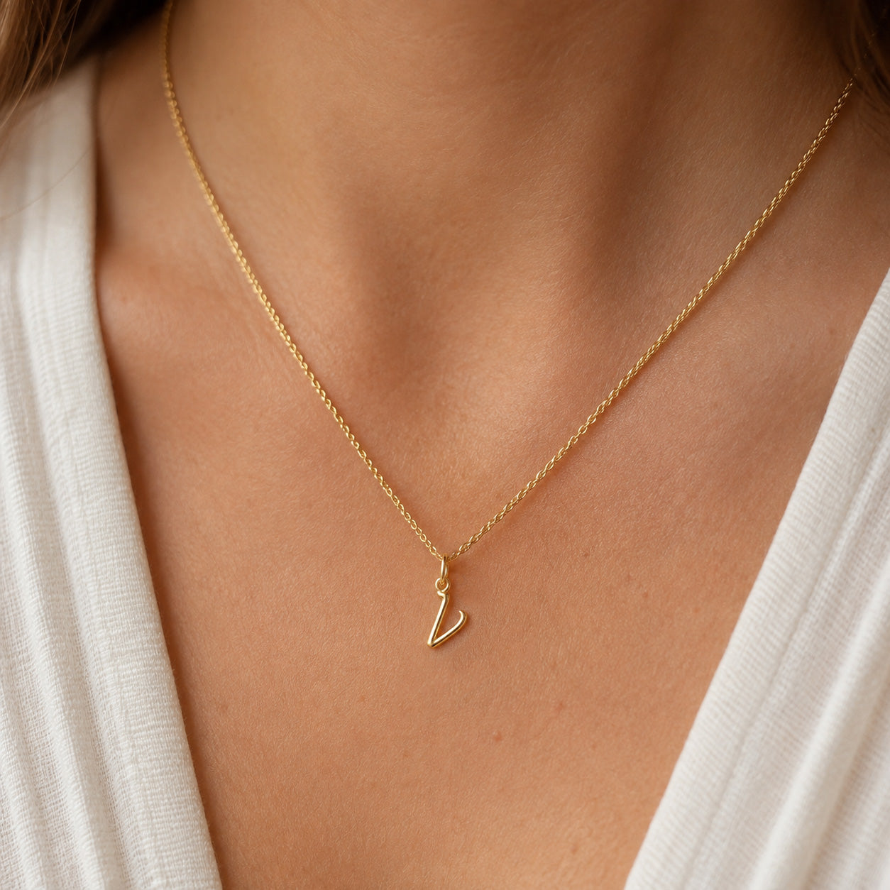 [group: v-10k-gold]A close-up of a person's neck and chest wearing a delicate gold chain necklace with a small, openwork initial 'L' pendant, styled with a white top.