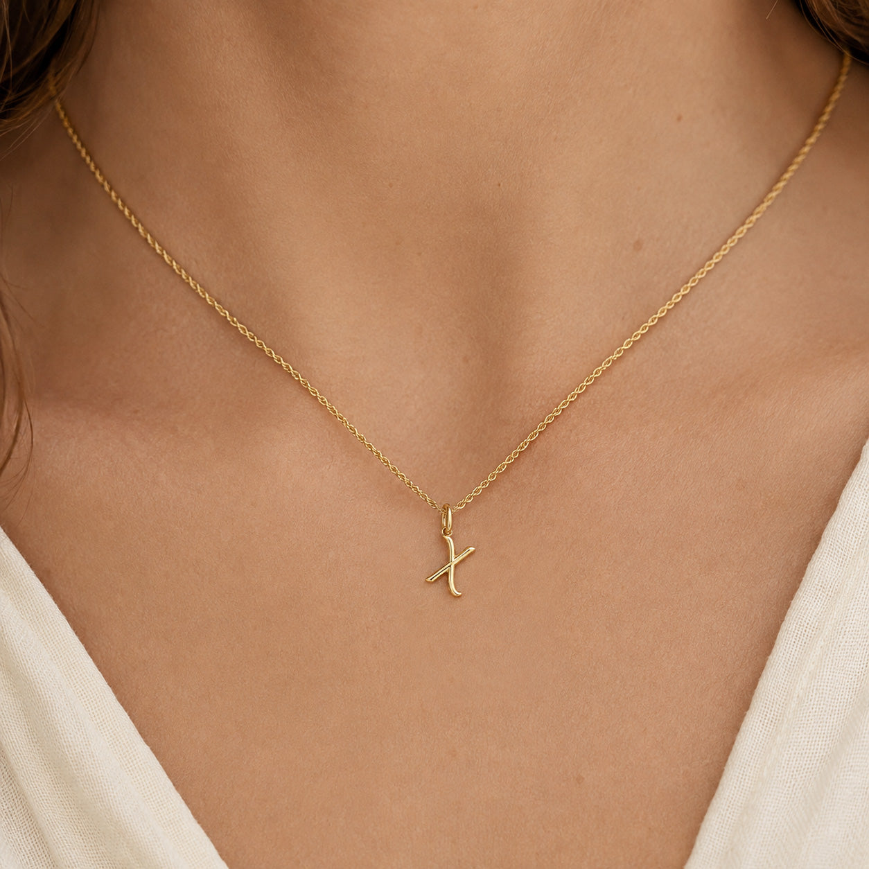 [group: x-10k-gold]A close-up of a model wearing a delicate gold chain necklace with a small, stylized gold pendant on a cream-colored top.