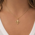 A close-up of a model wearing a delicate gold chain necklace with a small fleur-de-lis pendant, styled with a white ribbed top.