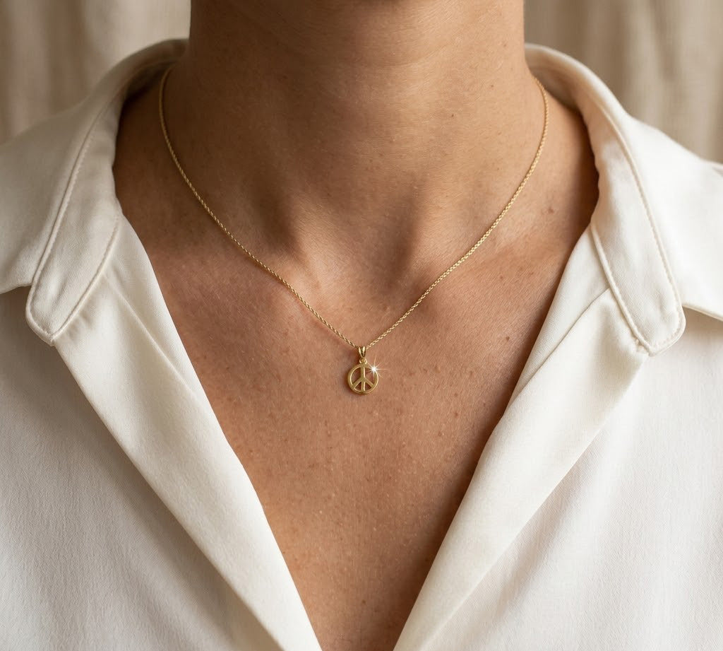A close-up of a person wearing a delicate gold chain necklace with a small peace sign pendant, styled with an off-white collared shirt.
