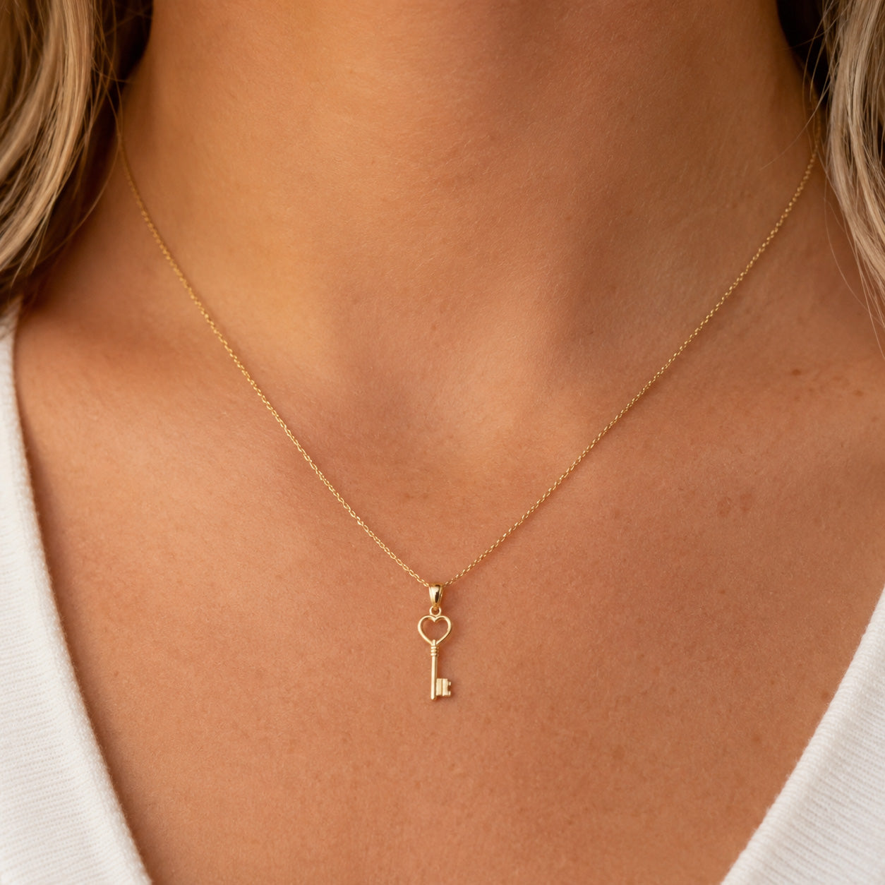 A delicate gold key pendant necklace with a heart-shaped key bow, worn on a model's neck over a white top.