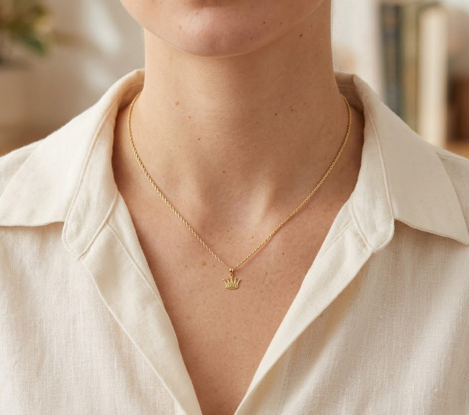 [group: small]A close-up of a person wearing a delicate gold chain necklace with a small crown pendant, styled with a cream-colored linen shirt.