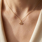 [group: yellow]A delicate gold crown pendant necklace worn on a model's neck, framed by soft cream-colored clothing under warm, direct sunlight.