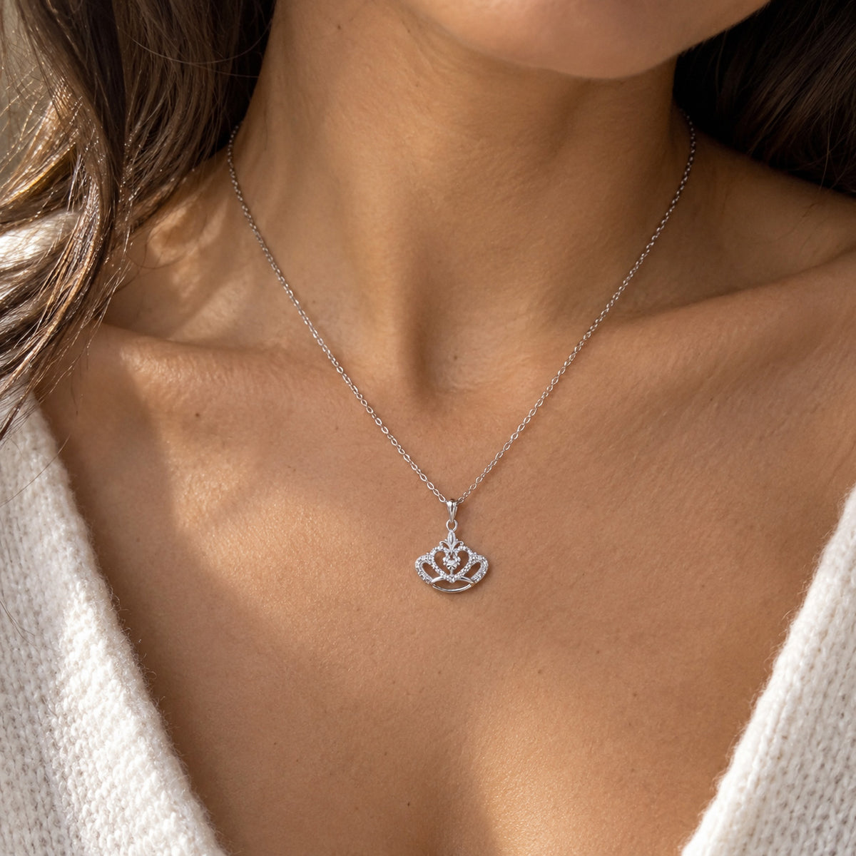 A close-up of a woman's neck and chest wearing a delicate silver chain necklace with a small crown-shaped pendant set with clear stones.