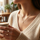 A close-up lifestyle shot of a woman wearing a delicate gold initial necklace with the letter 'A' pendant, holding a cup of coffee in a warm, softly blurred cafe setting.