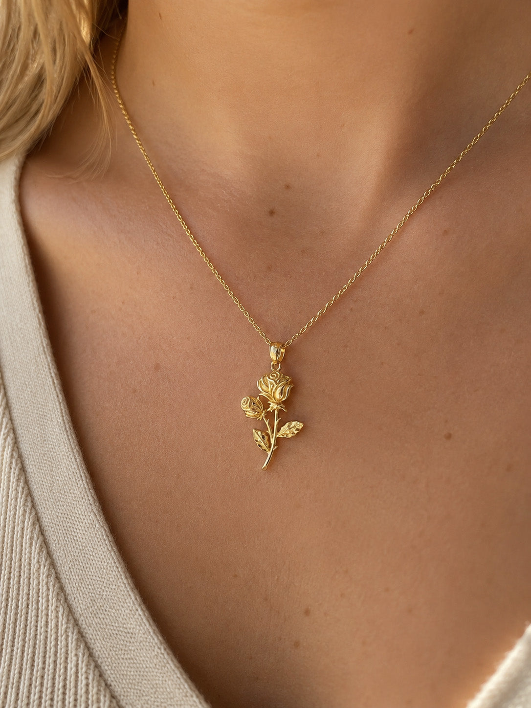 A close-up of a model wearing a delicate gold necklace with a rose-shaped pendant, styled with a beige knit top.