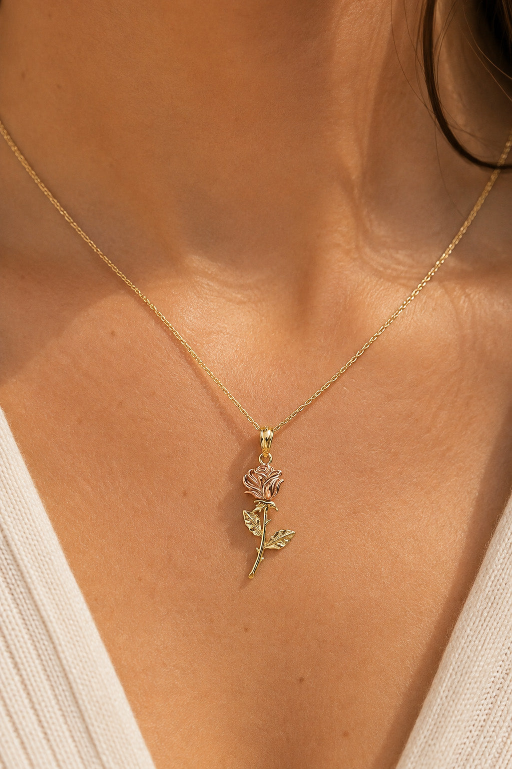 A close-up of a model's neck and chest wearing a delicate gold chain necklace with a rose-shaped pendant featuring pink and green enamel details.