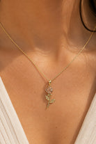 A close-up of a model's neck and chest wearing a delicate gold chain necklace with a rose-shaped pendant featuring pink and green enamel details.