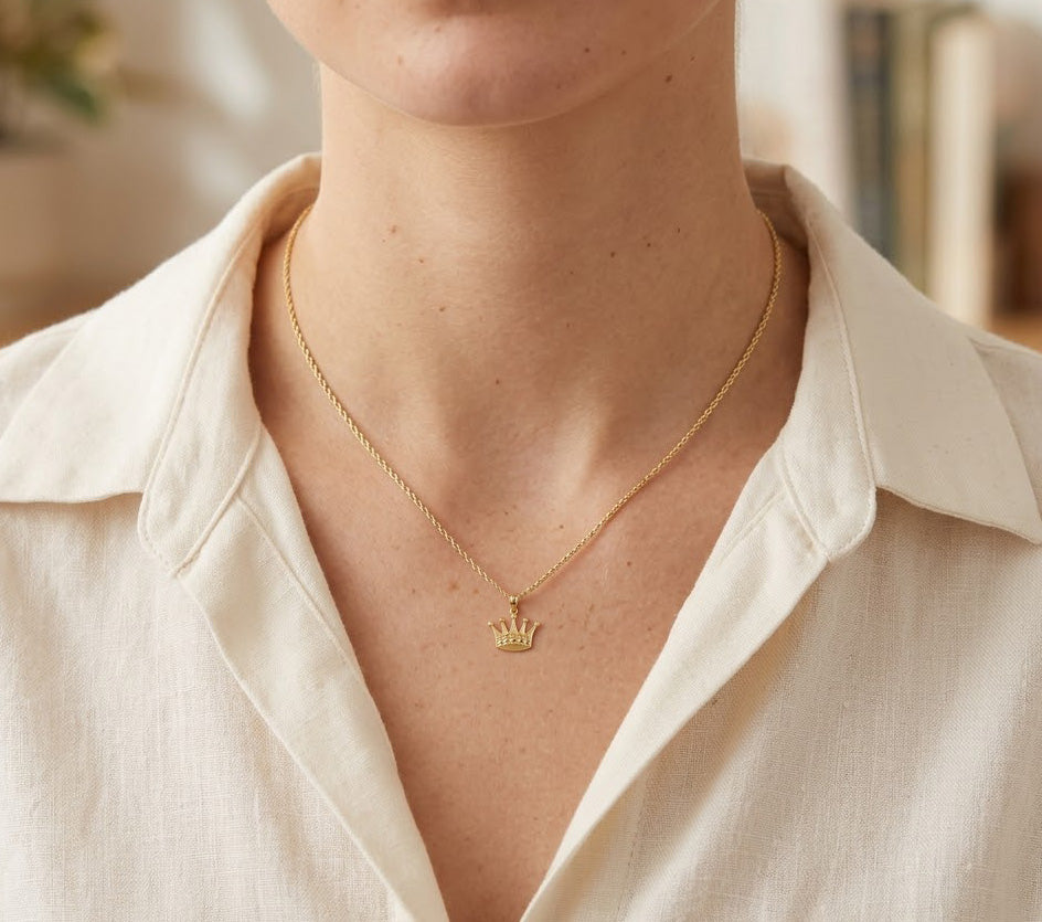 [group: large]A close-up of a person wearing a delicate gold chain necklace with a small crown pendant, styled with a cream-colored collared shirt.