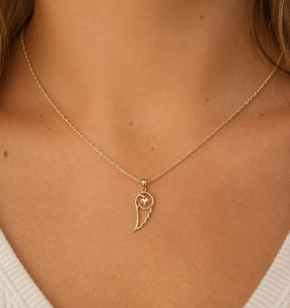 A delicate gold chain necklace with a wing-shaped pendant featuring a small pink heart in the center, worn on a model's neck.