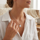 [group: silver]A close-up lifestyle shot of a woman wearing a delicate silver chain necklace with a round, textured silver pendant, styled with a white linen shirt in a warm, softly lit indoor setting.