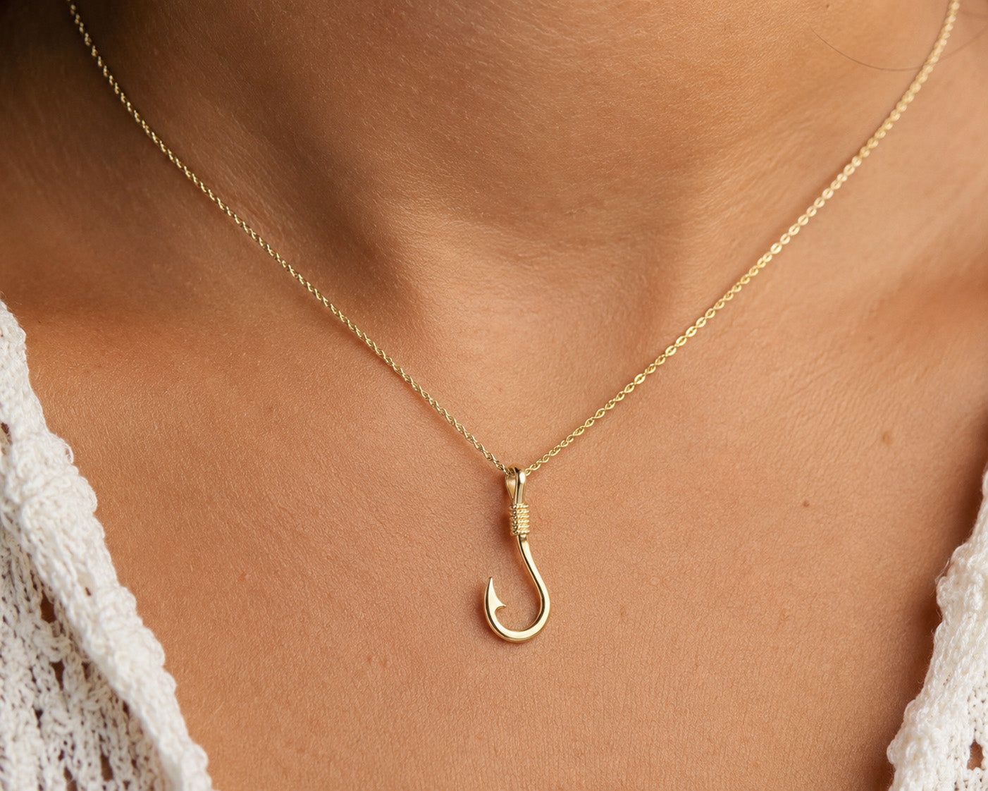 A gold necklace with a hook-shaped pendant worn on a person's neck, shown in a close-up shot with a white lace top visible at the edges.