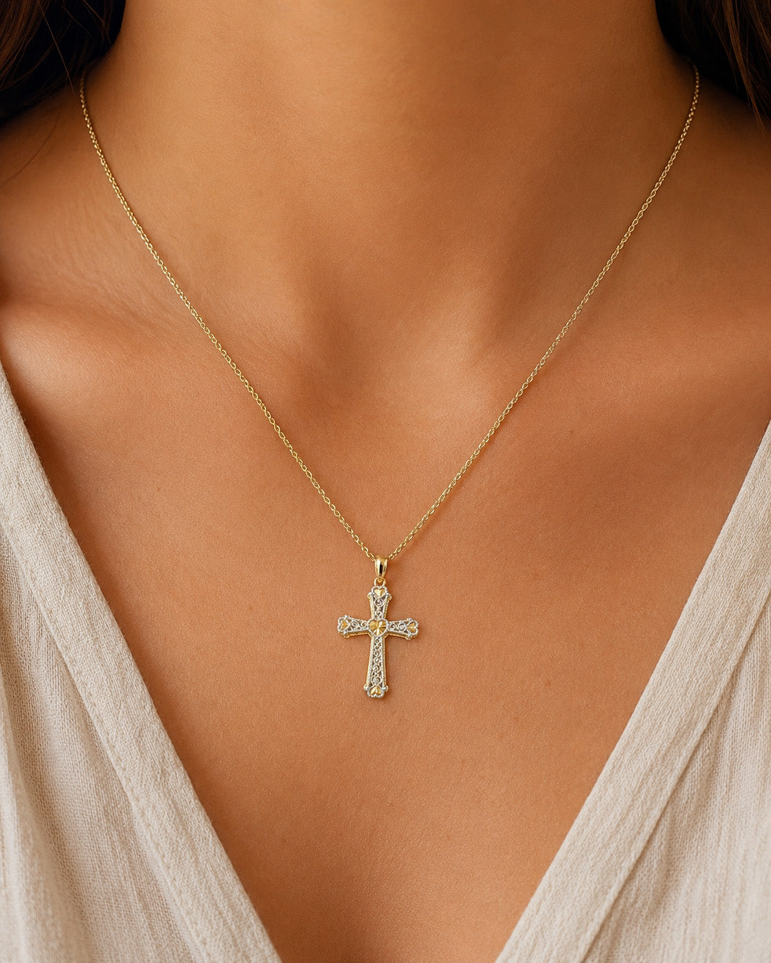[group: 14k-gold]A close-up of a model's neck and chest wearing a delicate gold cross pendant necklace adorned with small clear stones, paired with a light beige V-neck top.