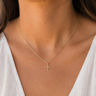 A delicate gold chain necklace with a small cross pendant, worn by a model in a white top.