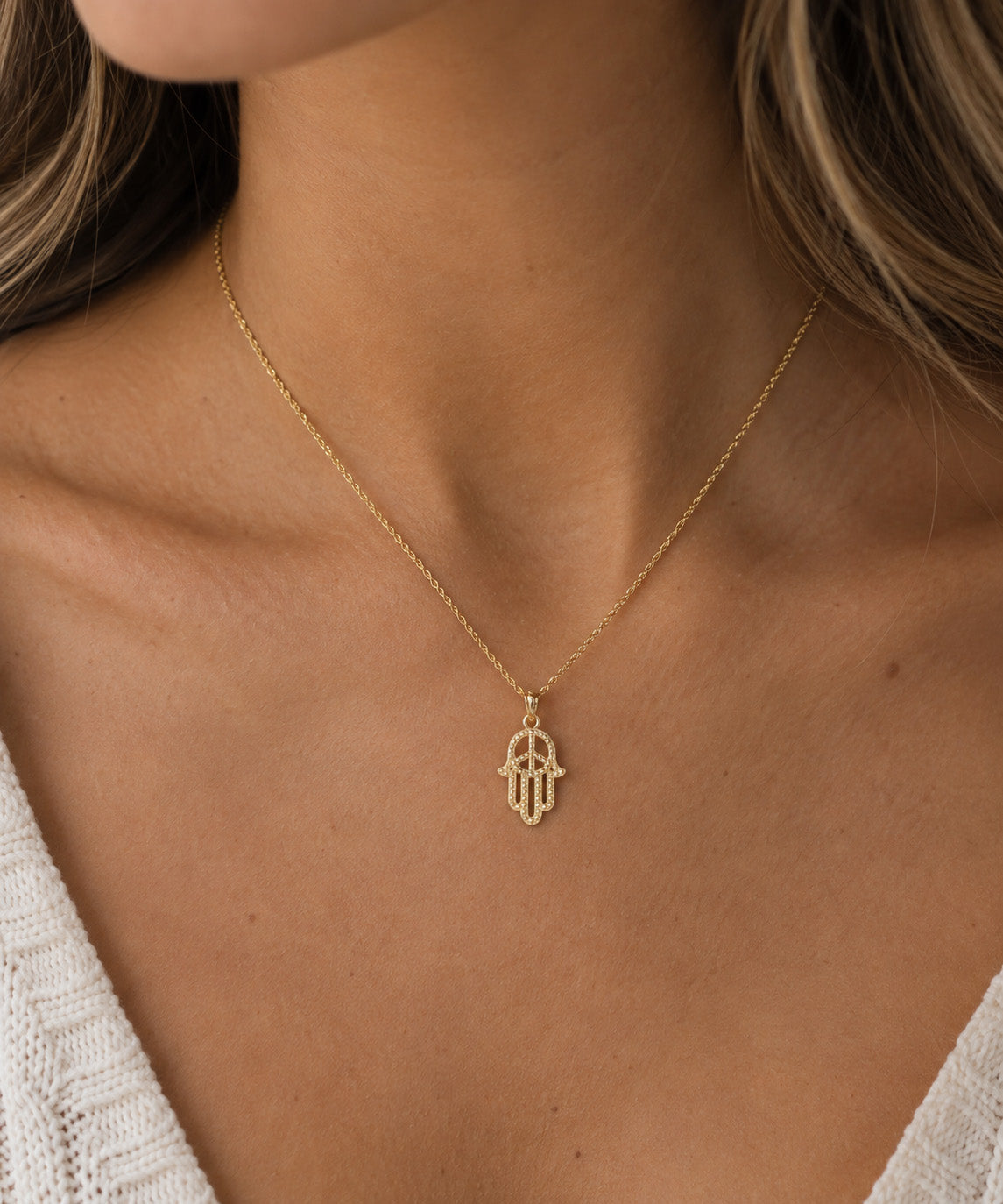 A close-up of a model wearing a delicate gold chain necklace with a Hamsa hand pendant, styled with a white knit top.