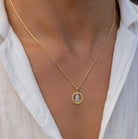 A close-up of a person wearing a delicate gold chain necklace with a circular pendant featuring a small white crystal or gemstone figure, styled with a white collared shirt.