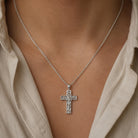 A silver cross pendant necklace with an engraved design, worn on a model's neck over a light beige button-down shirt.