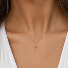 A delicate gold cross pendant necklace worn on a model's neck, shown in a close-up shot against a white V-neck top.