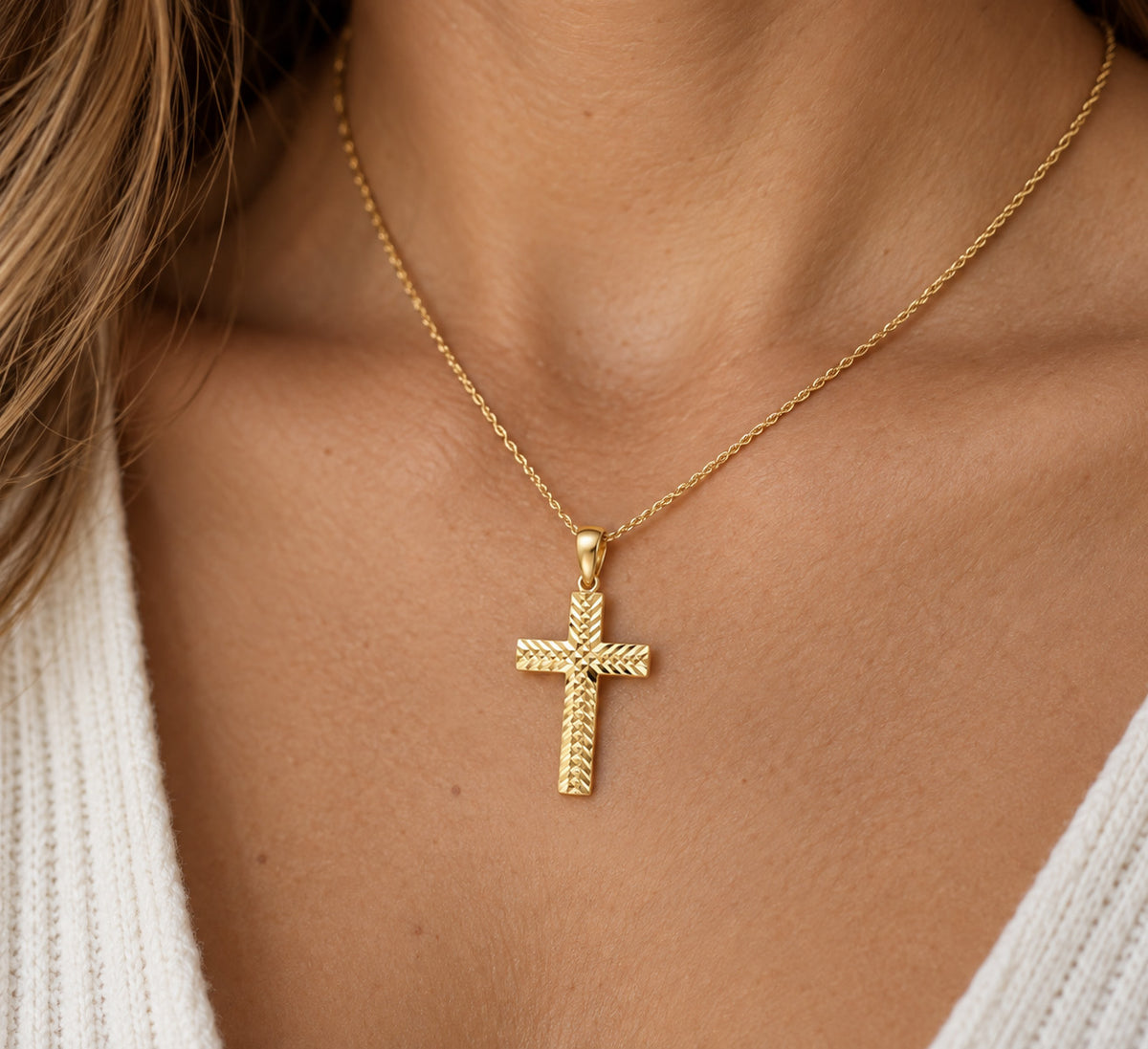 A gold cross pendant necklace with a textured rope-style finish, worn on a model's neck over a white knit top.