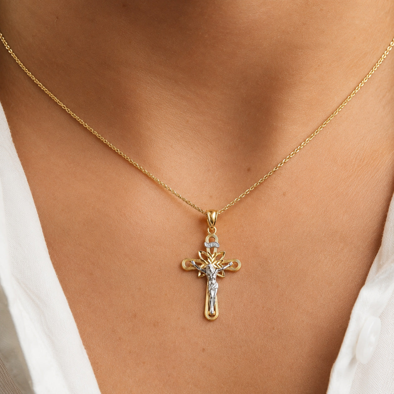 A close-up of a person's neck and chest wearing a delicate gold chain necklace with a cross pendant. The pendant features a gold figure with a small light blue stone at the top and a textured gold background.