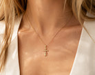 A close-up of a model wearing a delicate gold cross pendant necklace on a fine chain, styled with a white ribbed top.