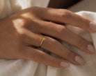 A delicate gold ring with a small cross detail worn on a person's ring finger, resting on soft white fabric.