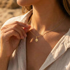 Close-up of a person wearing a gold necklace with a pendant, set against a blurred natural background.