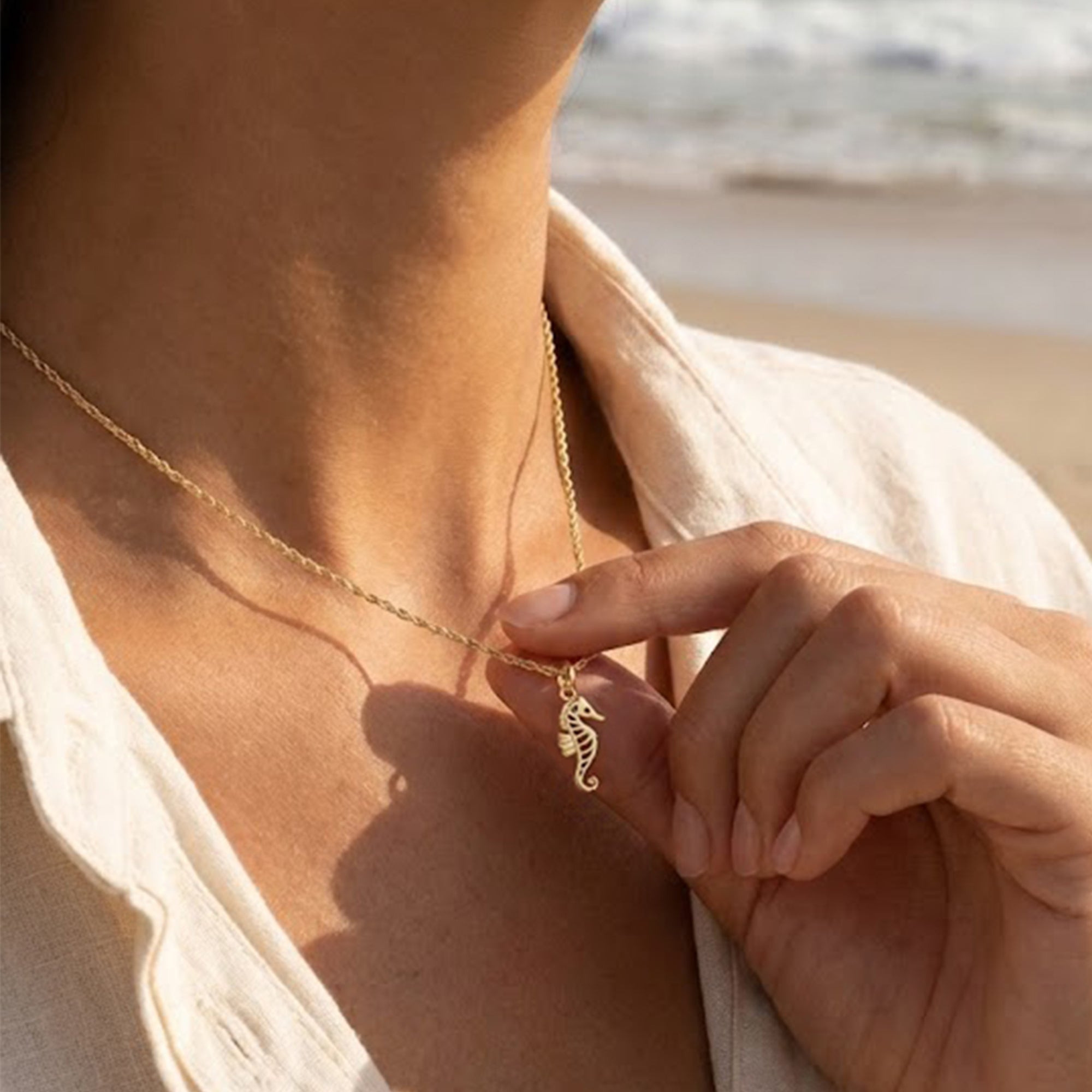 Person wearing a gold necklace with a seahorse pendant on a beach.